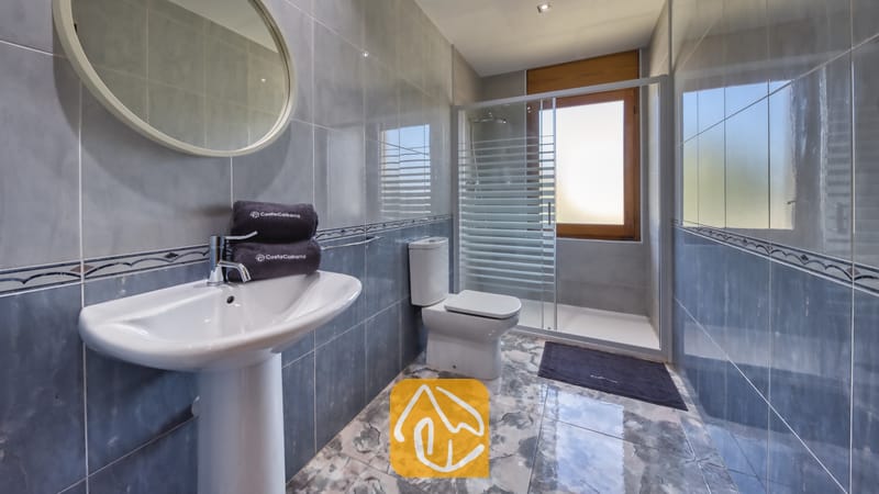 Betegelde badkamer met een wastafel op zuil, een ronde spiegel, een toilet en een glazen doucheruimte met een matglas raam.