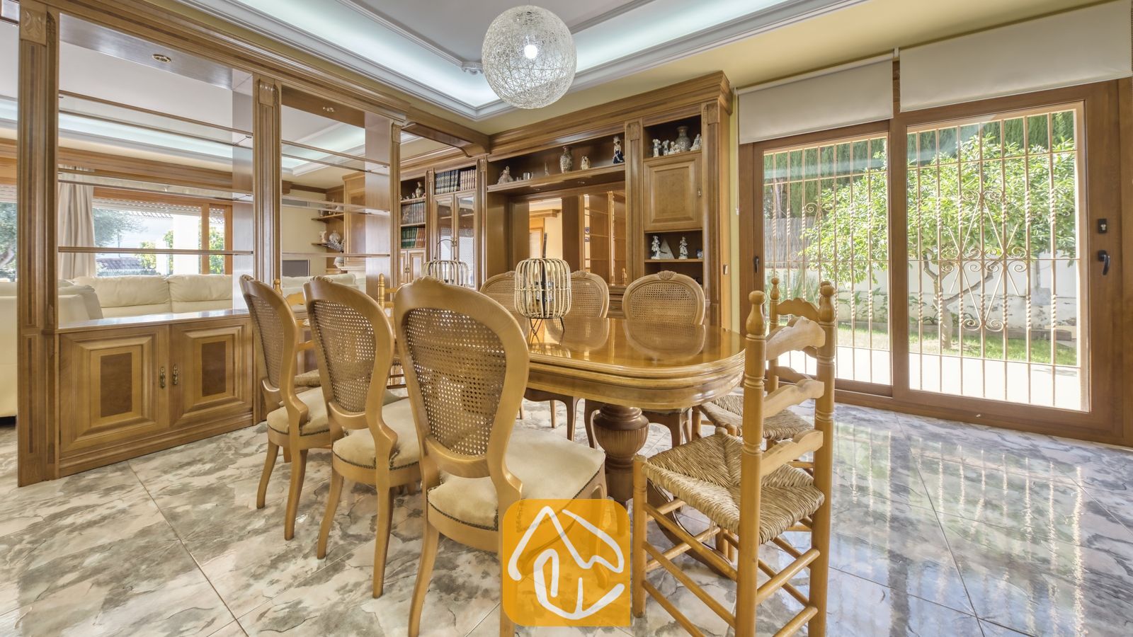 Eetkamer met een gepolijste houten tafel, stoelen met rieten rugleuningen, inbouwkasten van hout, marmeren vloeren en grote glazen schuifpui die uitzicht biedt op een tuin.