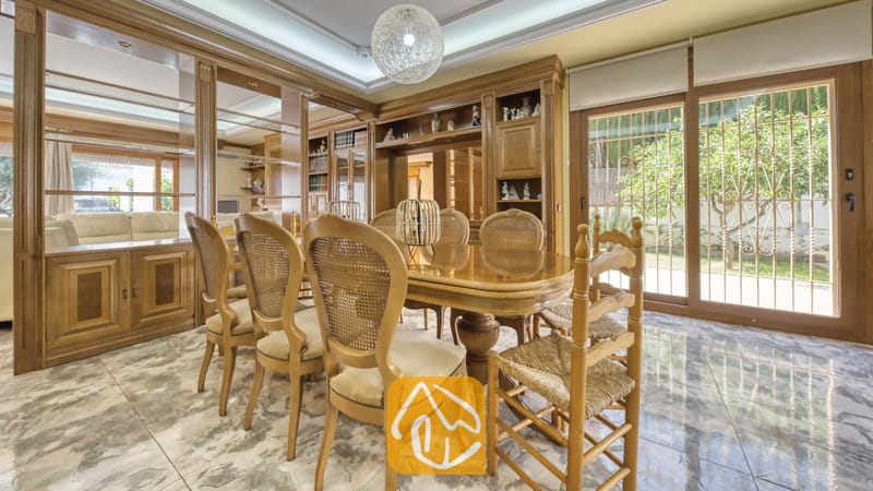 Eetkamer met een gepolijste houten tafel, stoelen met rieten rugleuningen, inbouwkasten van hout, marmeren vloeren en grote glazen schuifpui die uitzicht biedt op een tuin.