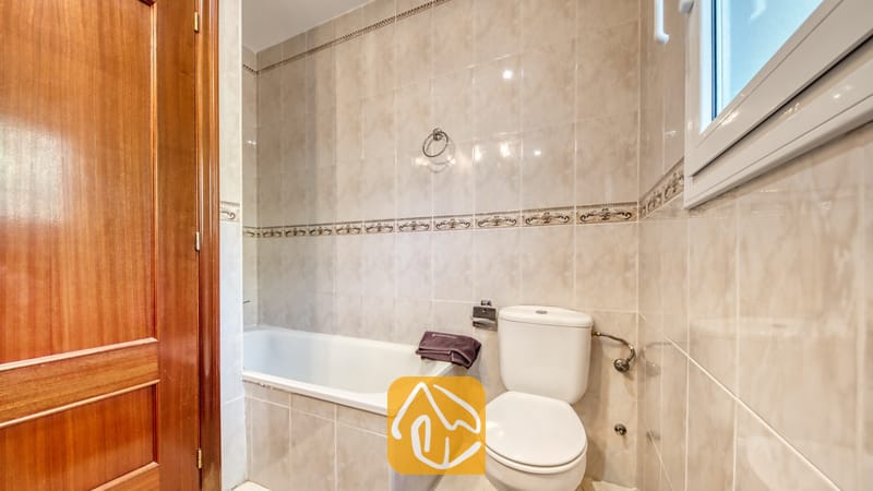 Badkamer voorzien van een wit porseleinen ligbad en toilet, neutrale beige tegels met een decoratieve rand en een gepolijste houten deur.