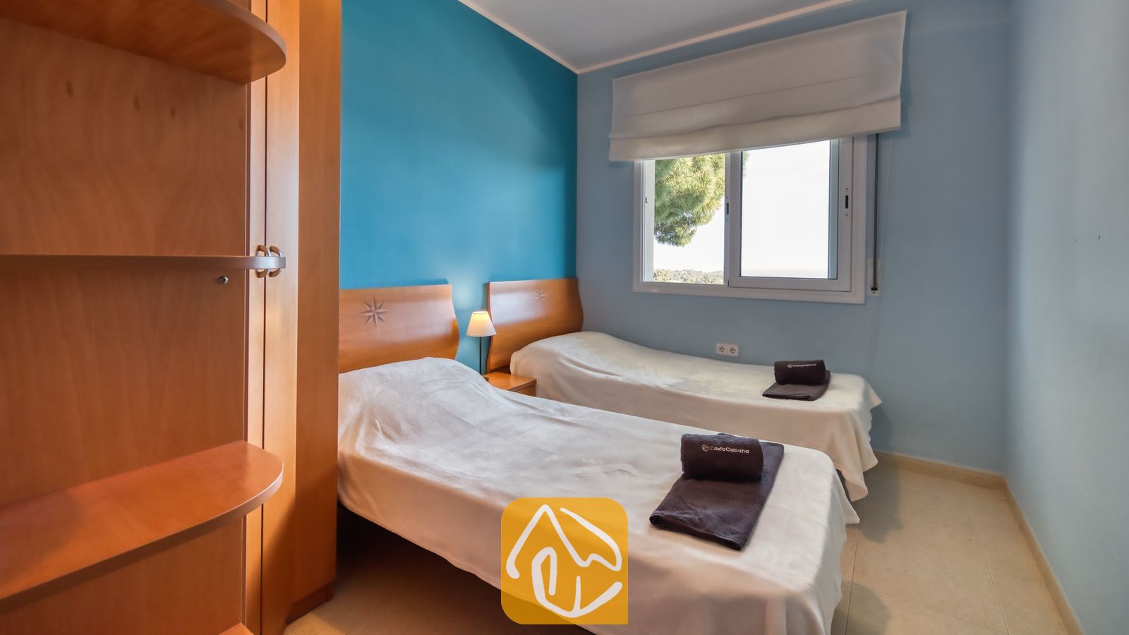 Tweepersoonskamer in Villa Chanel, voorzien van twee eenpersoonsbedden met wit bedlinnen, houten hoofdborden en een blauwe accentmuur naast een raam met witte jaloezieën.