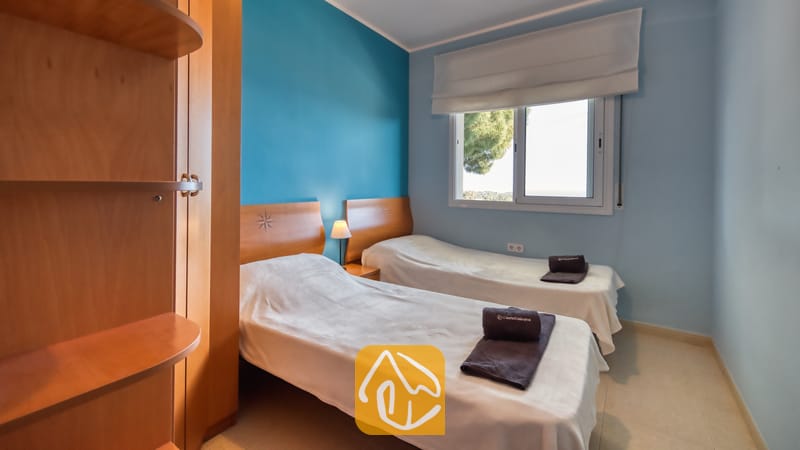 Tweepersoonskamer in Villa Chanel, voorzien van twee eenpersoonsbedden met wit bedlinnen, houten hoofdborden en een blauwe accentmuur naast een raam met witte jaloezieën.