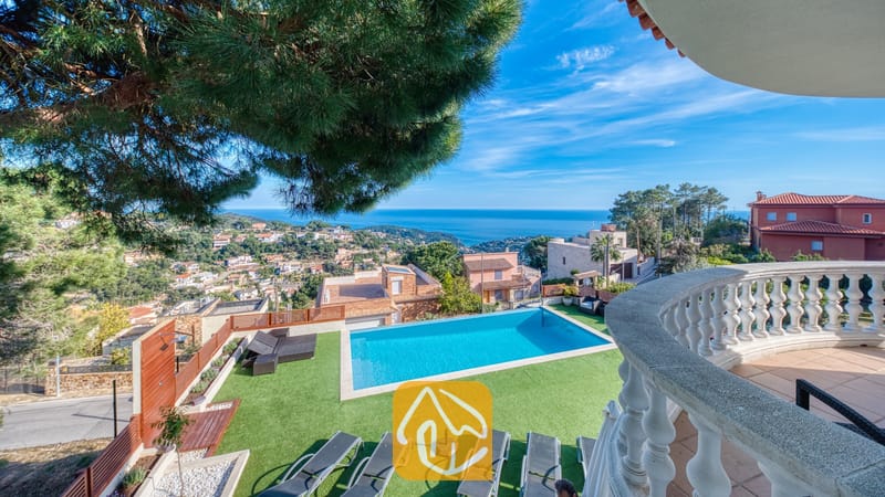 Privézwembad bij Villa Chanel met grijze ligbedden op een groen gazon, gezien vanaf een stenen balkon met uitzicht op de zee en de heuvels langs de kust.
