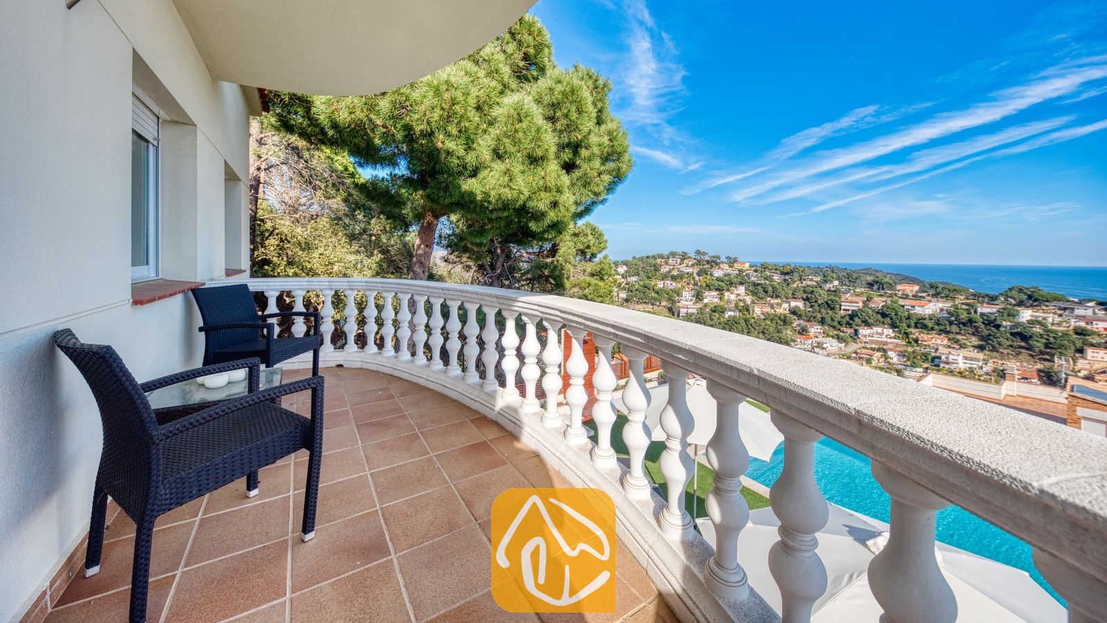 Gebogen balkon bij Villa Chanel, voorzien van zwart vlechtwerkmeubilair, een witstenen balustrade en uitzicht op de kustplaats en de zee.