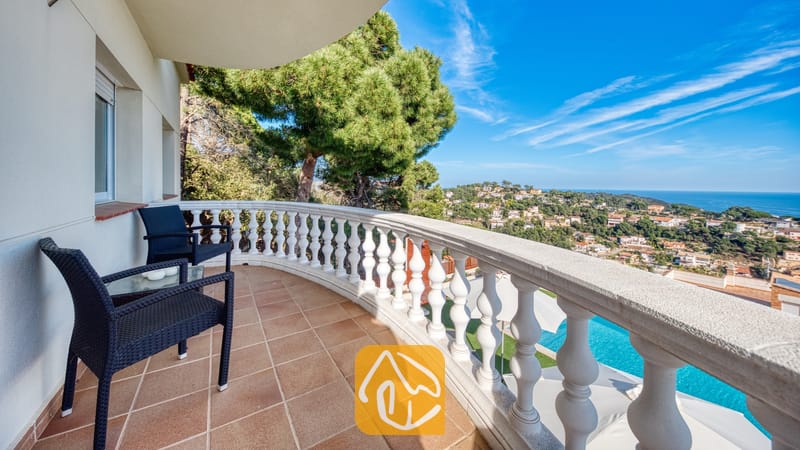 Gebogen balkon bij Villa Chanel, voorzien van zwart vlechtwerkmeubilair, een witstenen balustrade en uitzicht op de kustplaats en de zee.