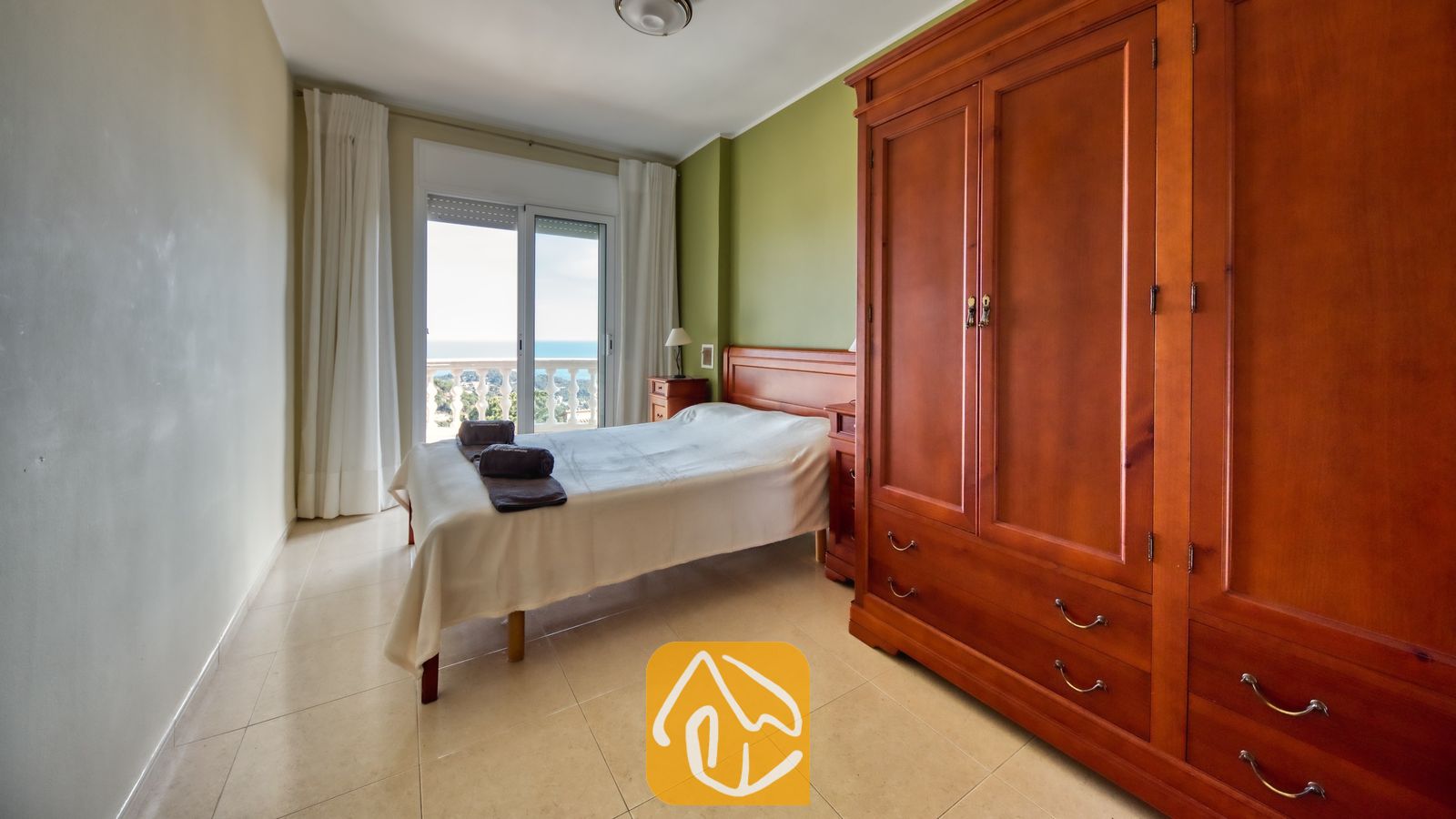 Slaapkamer met een grote houten kledingkast, een groene accentmuur en een bed met wit bedlinnen. Glazen deuren bieden toegang tot een balkon met uitzicht op zee bij Villa Chanel.