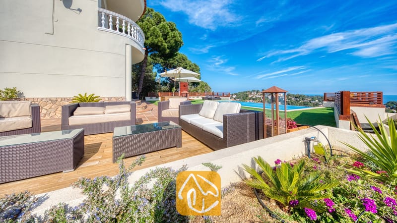 Terraslounge bij Villa Chanel, voorzien van donker vlechtwerkmeubilair met crèmekleurige kussens, glazen tafels en een infinity pool met uitzicht op zee.