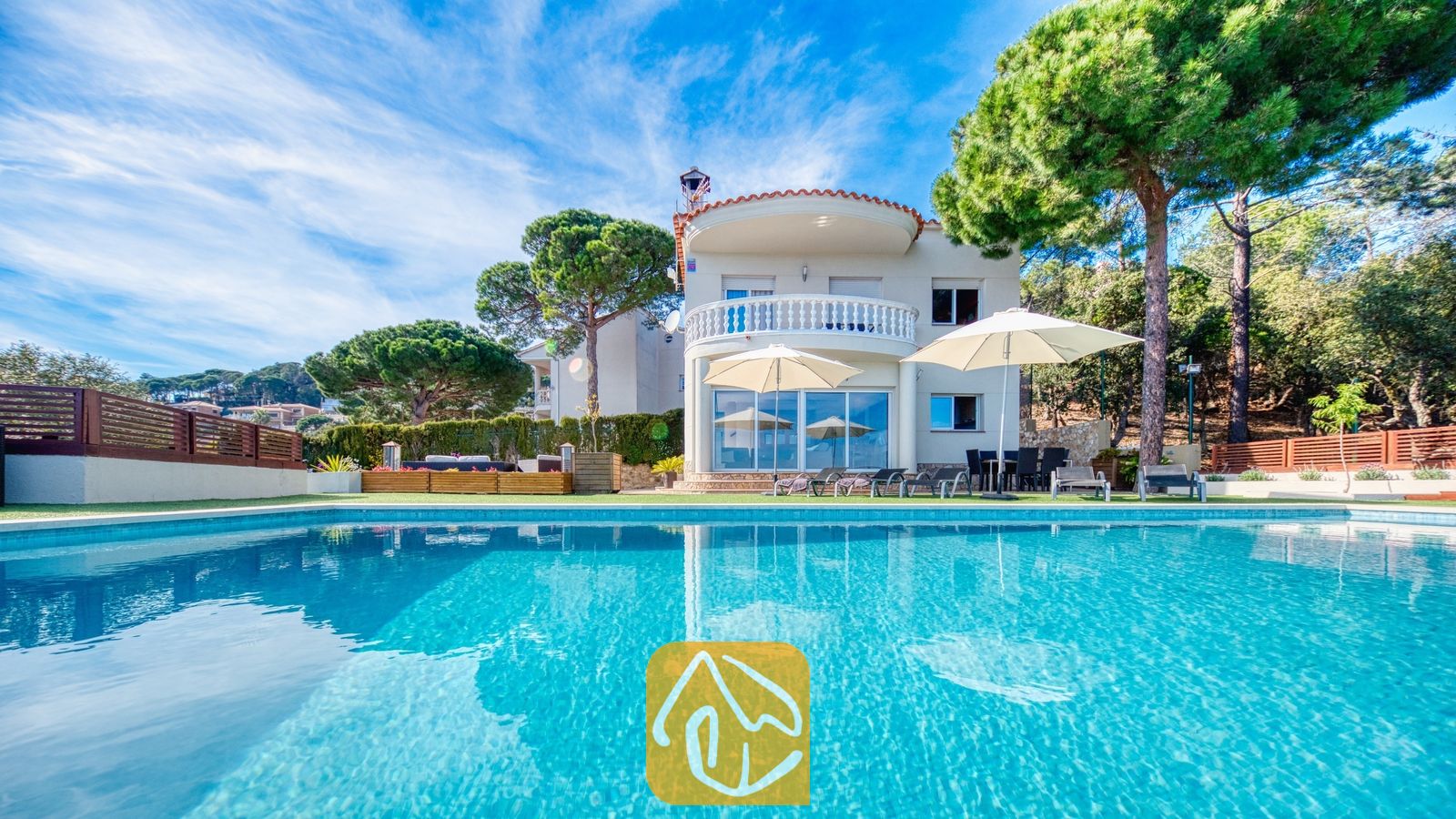 De buitenkant van Villa Chanel met een turquoise zwembad, witte parasols op het terras en een gebogen stenen balkon, omringd door weelderige groene dennenbomen.