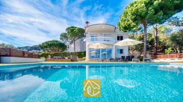 De buitenkant van Villa Chanel met een turquoise zwembad, witte parasols op het terras en een gebogen stenen balkon, omringd door weelderige groene dennenbomen.