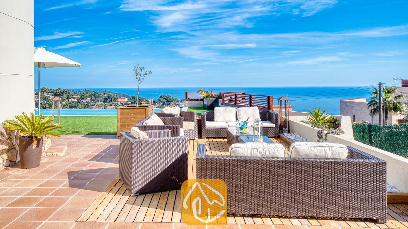Terras van Villa Chanel met bruin vlechtwerk loungeset, witte kussens en een houten vlonder met uitzicht op zee en een infinity pool.