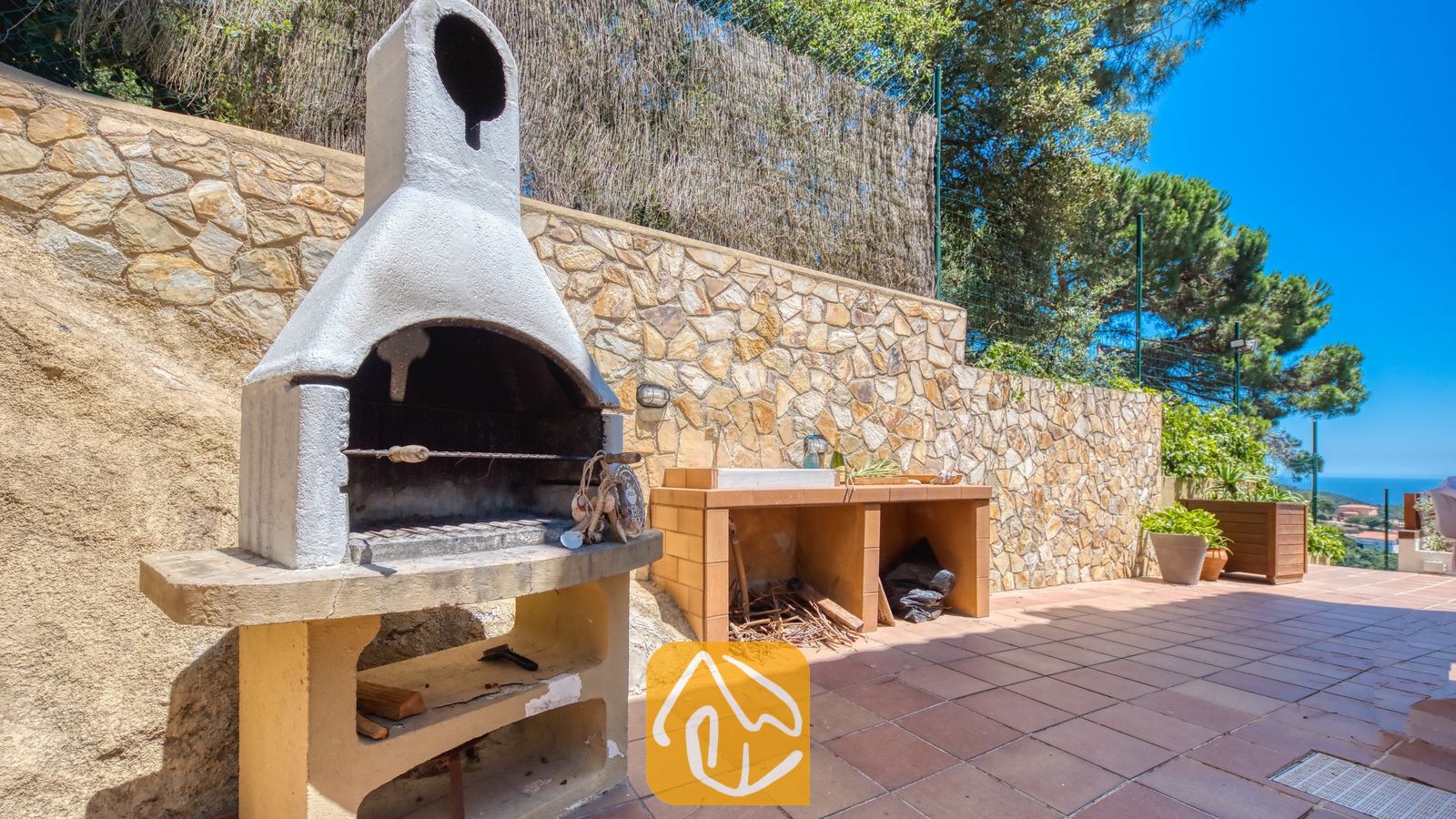 Buitenbarbecue van natuursteen en een spoelbak op een betegeld terras bij Villa Chanel, gekenmerkt door een stenen muur en een prachtig uitzicht op de verre zee.