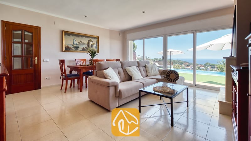 Woonkamer met een beige bank en een glazen salontafel in Villa Chanel, voorzien van schuifdeuren die toegang bieden tot een terras met zwembad en uitzicht op zee.