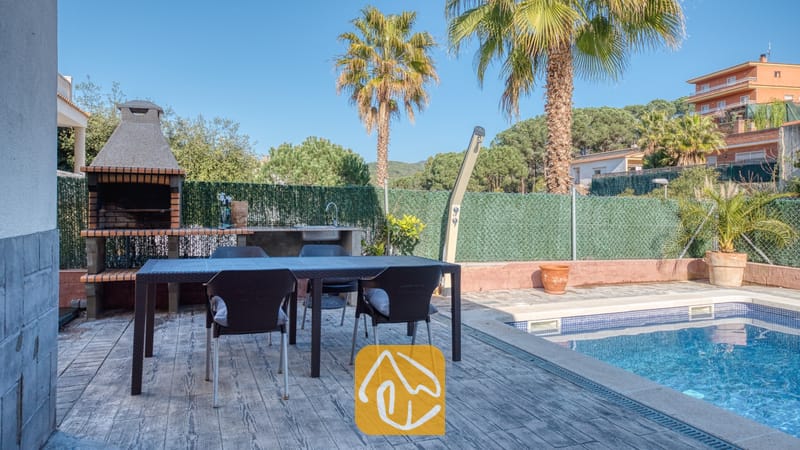 Eetgedeelte buiten bij het zwembad van Villa Zarita, voorzien van een donkere Wicker tuintafel, vier stoelen, een ingebouwde stenen barbecue en palmbomen onder een blauwe lucht.