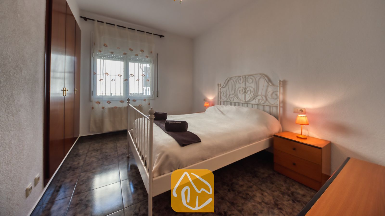 Slaapkamer met een wit metalen bed, donkere tegelvloer, houten nachtkastje met een oranje lamp en een inbouwkast.