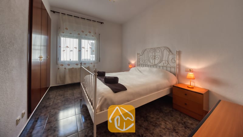 Slaapkamer met een wit metalen bed, donkere tegelvloer, houten nachtkastje met een oranje lamp en een inbouwkast.