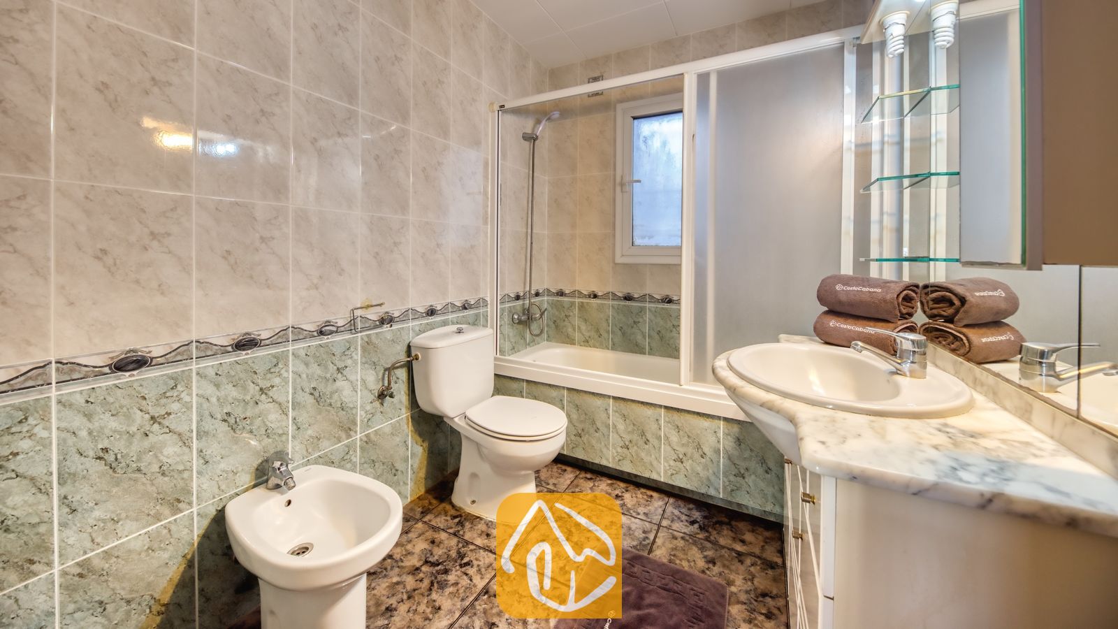Badkamer met muren van marmerlook tegels, een wit badmeubel met een waskom op het blad, een toilet, een bidet en een ligbad met glazen schuifdeuren.