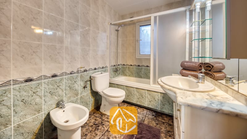Badkamer met muren van marmerlook tegels, een wit badmeubel met een waskom op het blad, een toilet, een bidet en een ligbad met glazen schuifdeuren.
