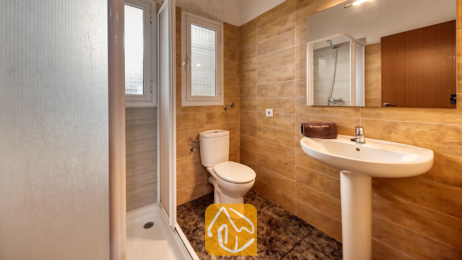 Badkamer met beige betegelde muren, een witte wastafel op zuil met een bruine handdoek, een toilet en een inloopdouche met glazen deur.