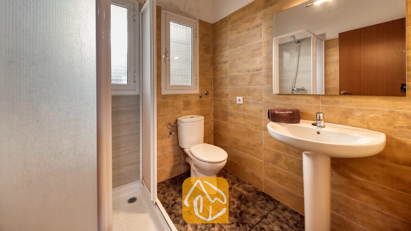 Badkamer met beige betegelde muren, een witte wastafel op zuil met een bruine handdoek, een toilet en een inloopdouche met glazen deur.