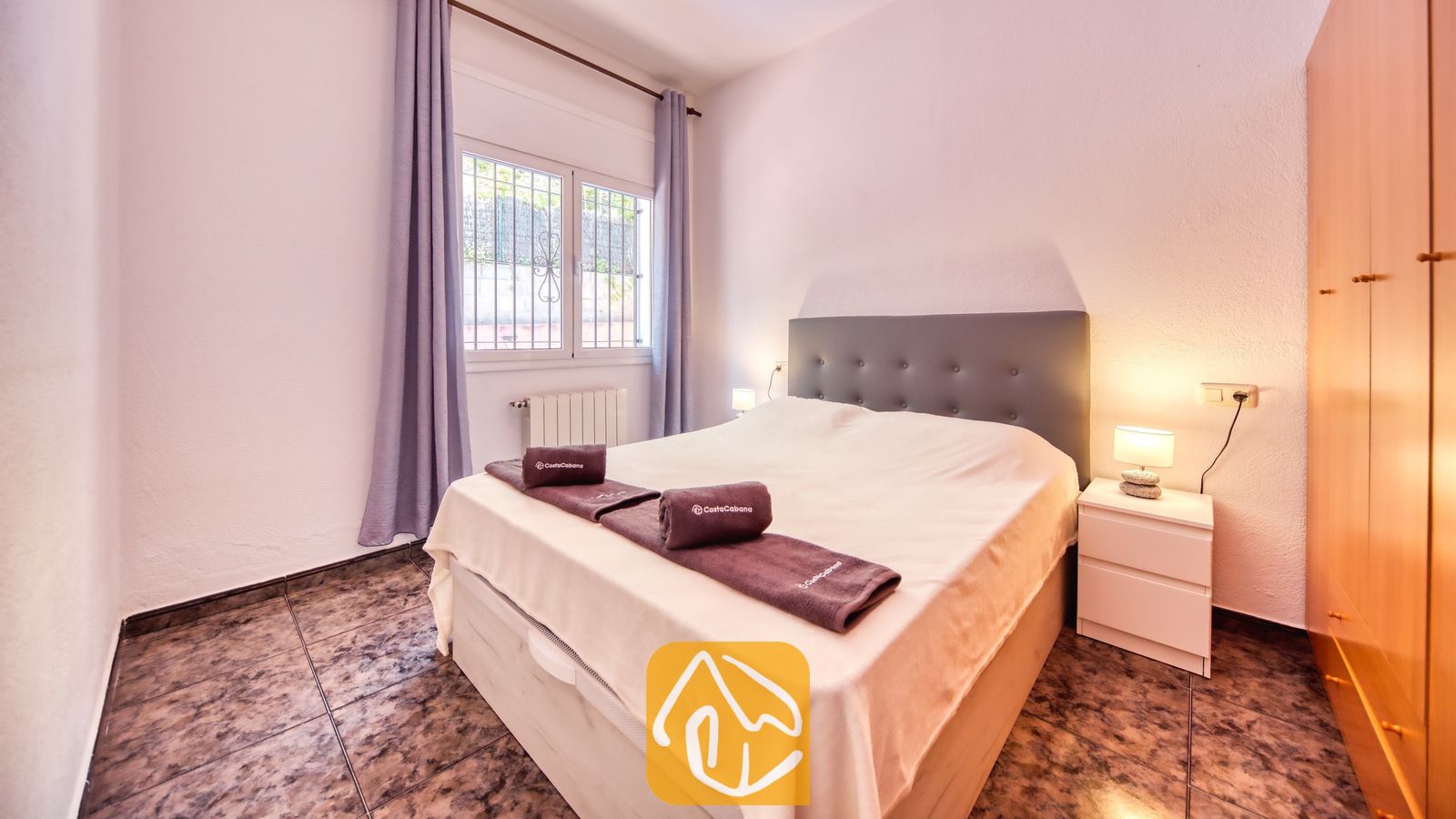 Slaapkamer met een tweepersoonsbed, een donkergrijze gestoffeerde hoofdbord, opgerolde handdoeken, witte nachtkastjes met lampen, paarse gordijnen en een tegelvloer.