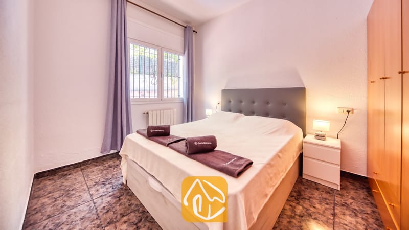 Slaapkamer met een tweepersoonsbed, een donkergrijze gestoffeerde hoofdbord, opgerolde handdoeken, witte nachtkastjes met lampen, paarse gordijnen en een tegelvloer.