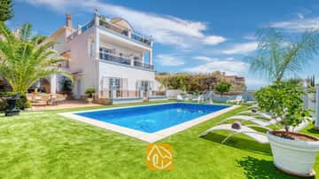 Privézwembad en witte ligbedden op een groen gazon bij Villa Maribel in Spanje, met een wit villa met meerdere verdiepingen, balkons en palmbomen.