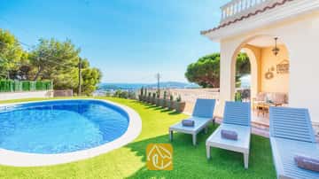 Privézwembad en blauwe houten ligstoelen op kunstgras bij Villa Ibiza met uitzicht op de verre zee en bomen.