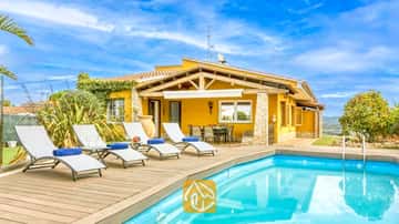Privézwembad met houten vlonder en witte ligstoelen bij Villa Soleia in Spanje, met een villa met een gele buitenkant en een blauwe lucht.